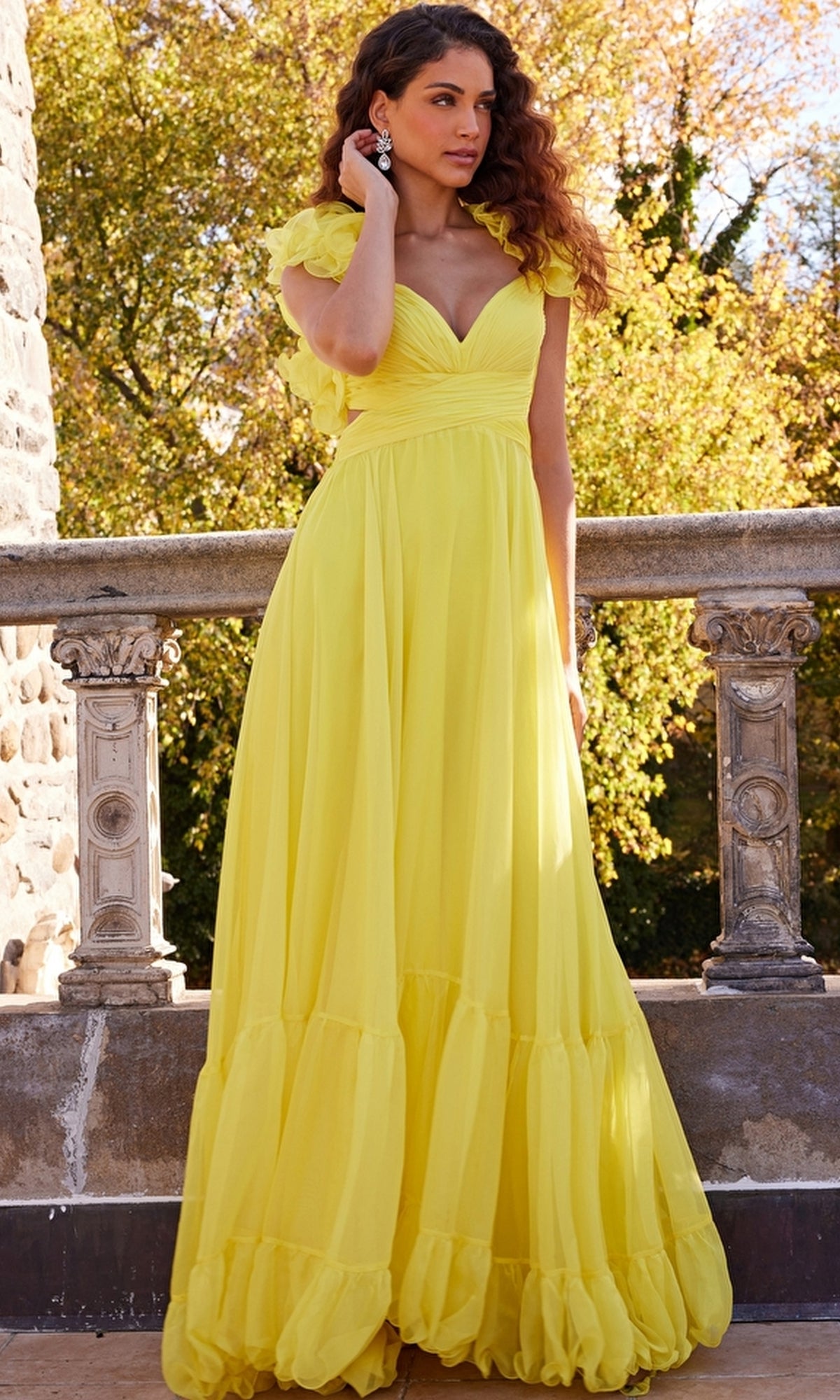 A woman stands outdoors on a stone balcony, wearing the Jovani Ruffled-Bodice Long Tiered Prom Dress 23322 with ruffled cap sleeves. Trees with autumn leaves are visible in the background.